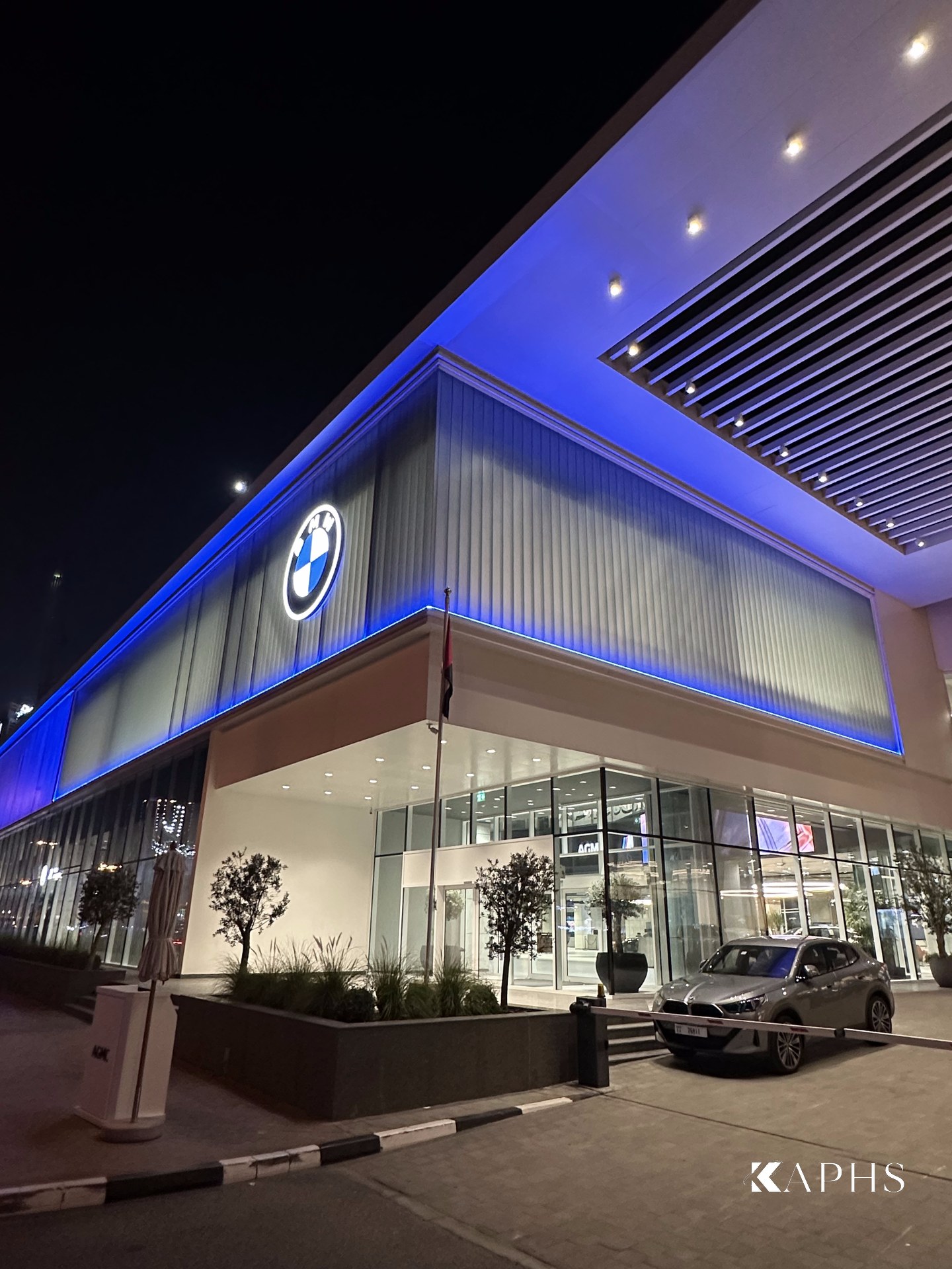 AGMC BMW showroom, Sheikh Zayed Road — corner view of the blue-lit U-channel glass façade and entrance canopy, KAPHS